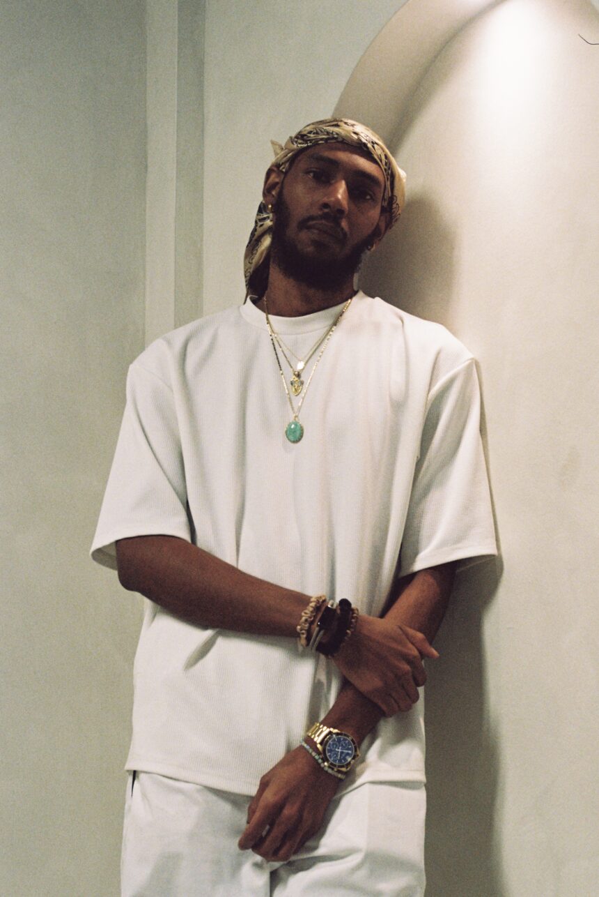 Keem wearing an all-white outfit, gold chain with green pendant, bandana headwrap, and a gold watch, leaning against an arched wall.