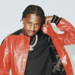 Lil Tjay poses in a red leather varsity jacket and gold chains against a light gray background.