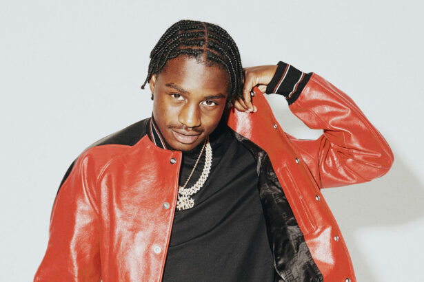 Lil Tjay poses in a red leather varsity jacket and gold chains against a light gray background.