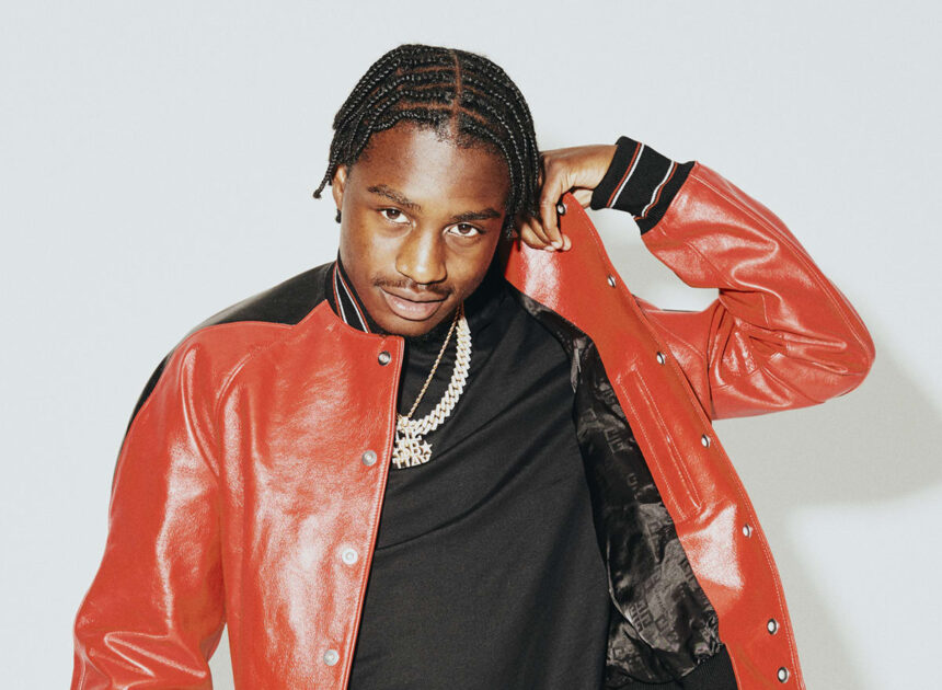 Lil Tjay poses in a red leather varsity jacket and gold chains against a light gray background.