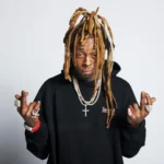 Lil Wayne posing in a black hoodie with diamond chains, rings, and his signature blonde dreadlock