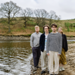 Manchester indie four-piece pyncher standing on a rocky riverbank surrounded by bare trees and rolling hills