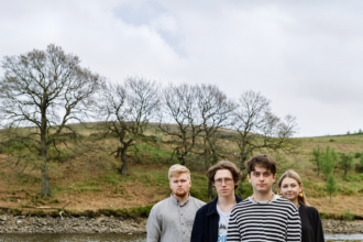 Manchester indie four-piece pyncher standing on a rocky riverbank surrounded by bare trees and rolling hills