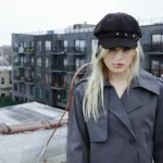 Musician underscores stands on a rooftop in a gray trench coat and black cap, with a city skyline behind her. Photo by Bailey Krawczyk.