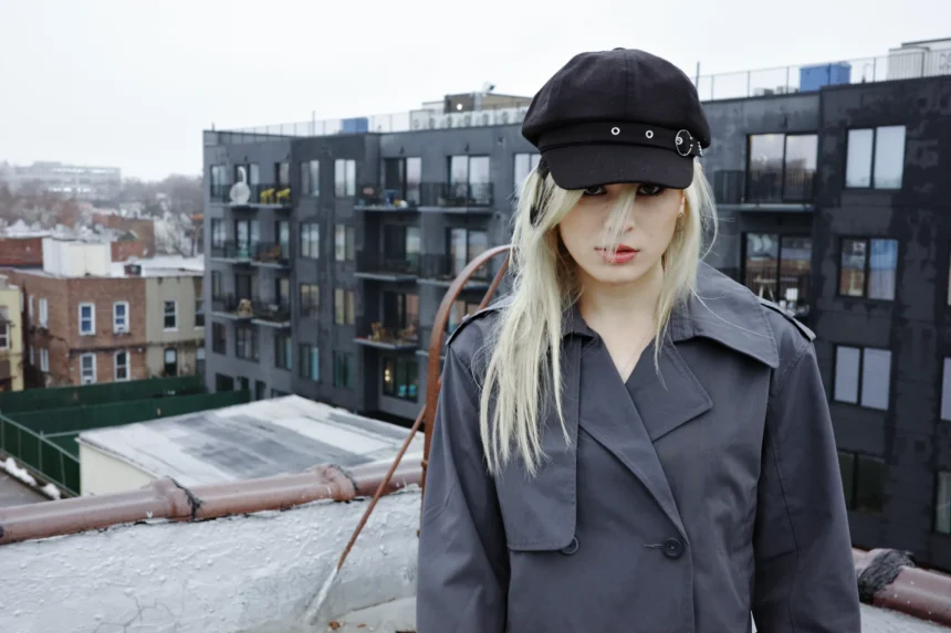 Musician underscores stands on a rooftop in a gray trench coat and black cap, with a city skyline behind her. Photo by Bailey Krawczyk.