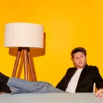 Niall Horan reclining casually at a table, wearing jeans and a black blazer, against a bright yellow background with a tripod floor lamp.
