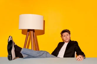 Niall Horan reclining casually at a table, wearing jeans and a black blazer, against a bright yellow background with a tripod floor lamp.
