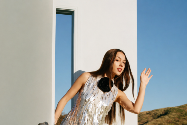 Olivia Rodrigo in a silver dress stepping through a white door in a sunny field LateTown.com