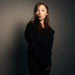 Portrait of Ariana Grande posing against a moody studio backdrop, wearing a black off-shoulder outfit with long brown hair and a playful expression, standing in soft dramatic lighting.