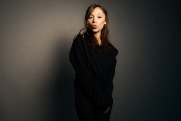 Portrait of Ariana Grande posing against a moody studio backdrop, wearing a black off-shoulder outfit with long brown hair and a playful expression, standing in soft dramatic lighting.