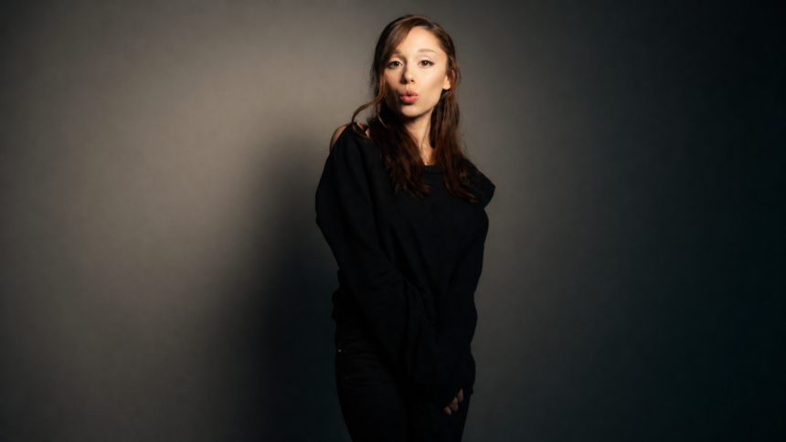 Portrait of Ariana Grande posing against a moody studio backdrop, wearing a black off-shoulder outfit with long brown hair and a playful expression, standing in soft dramatic lighting.