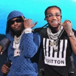 Offset and Quavo of Migos performing on stage, wearing diamond chains and designer outfits, holding microphones.