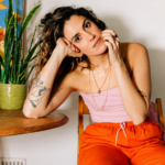 Shanti Celeste seated indoors beside a wooden table and potted plant, wearing a pink top and bright orange pants, looking thoughtfully at the camera in a warm, artistic setting.