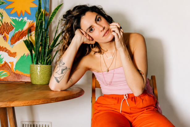 Shanti Celeste seated indoors beside a wooden table and potted plant, wearing a pink top and bright orange pants, looking thoughtfully at the camera in a warm, artistic setting.