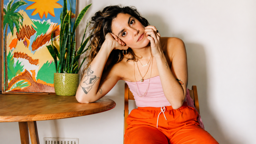 Shanti Celeste seated indoors beside a wooden table and potted plant, wearing a pink top and bright orange pants, looking thoughtfully at the camera in a warm, artistic setting.