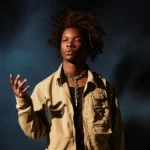 `Singer D4vd in a tan jacket and silver chain necklace against a dark blue background.`