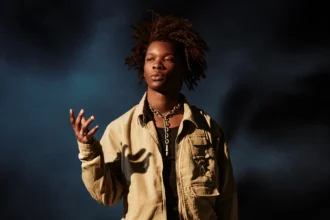 `Singer D4vd in a tan jacket and silver chain necklace against a dark blue background.`