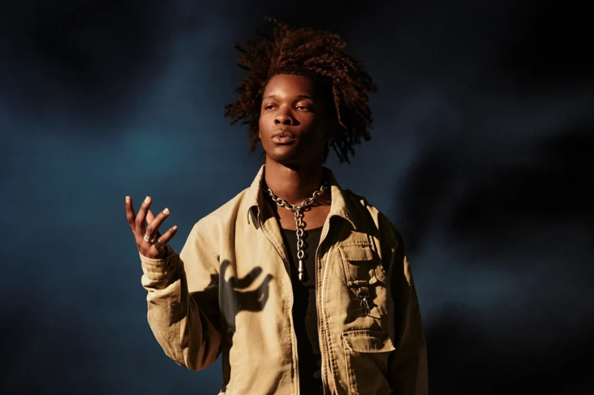 `Singer D4vd in a tan jacket and silver chain necklace against a dark blue background.`