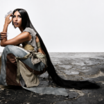 Surusinghe seated on cracked concrete, wearing layered earth tones with crystal accessories and floor-length black hair