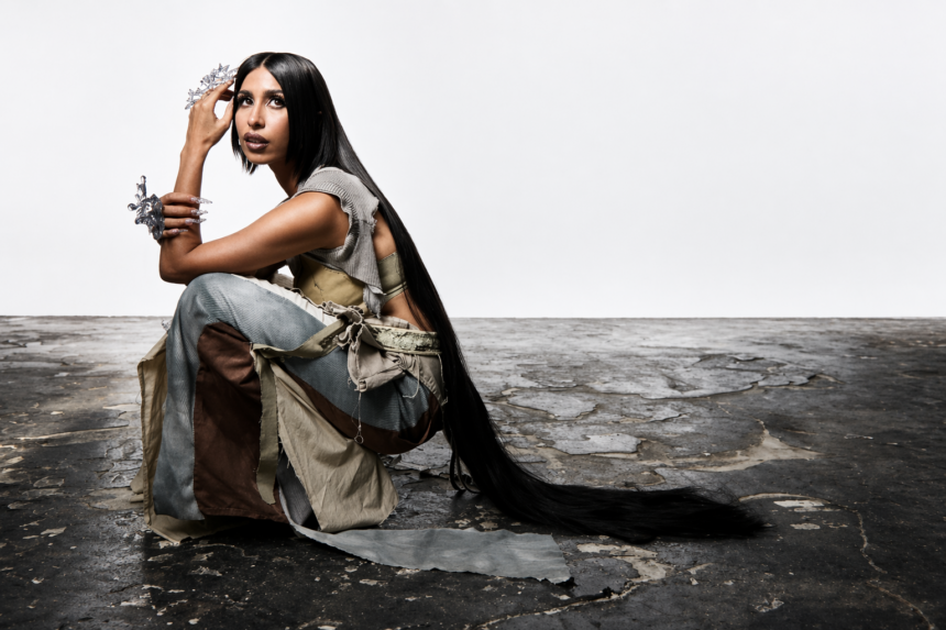 Surusinghe seated on cracked concrete, wearing layered earth tones with crystal accessories and floor-length black hair