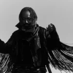 Thundercat poses in a black-and-white promo photo, wearing a fringed leather jacket with arms raised, head bowed, against a plain light background.