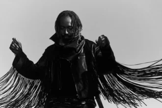 Thundercat poses in a black-and-white promo photo, wearing a fringed leather jacket with arms raised, head bowed, against a plain light background.