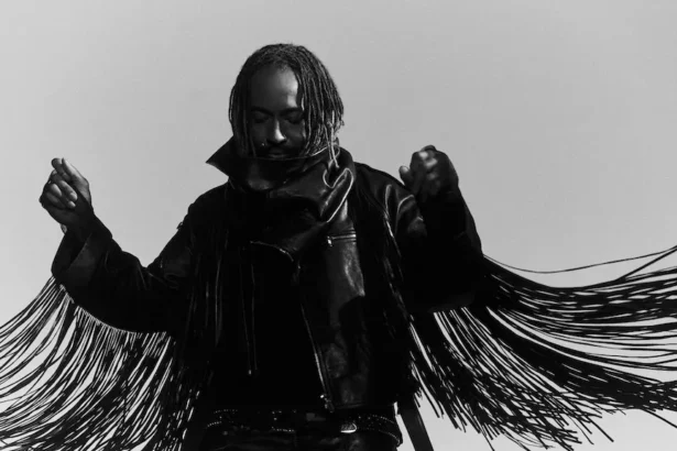 Thundercat poses in a black-and-white promo photo, wearing a fringed leather jacket with arms raised, head bowed, against a plain light background.