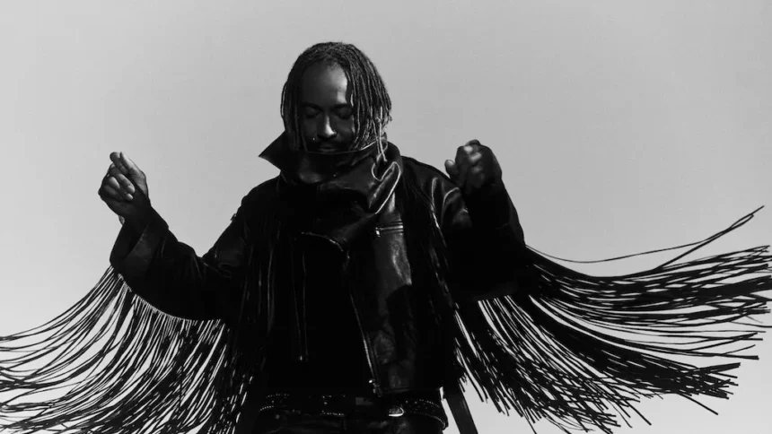 Thundercat poses in a black-and-white promo photo, wearing a fringed leather jacket with arms raised, head bowed, against a plain light background.