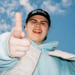 Venbee points playfully at the camera under a bright blue sky, wearing a black cap, light blue hoodie, and white jacket, capturing a candid, energetic moment.