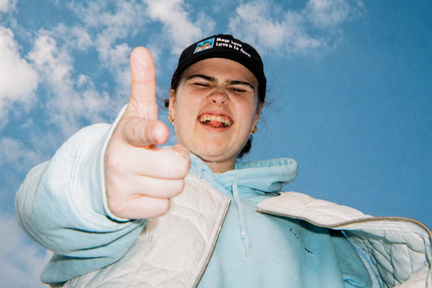 Venbee points playfully at the camera under a bright blue sky, wearing a black cap, light blue hoodie, and white jacket, capturing a candid, energetic moment.