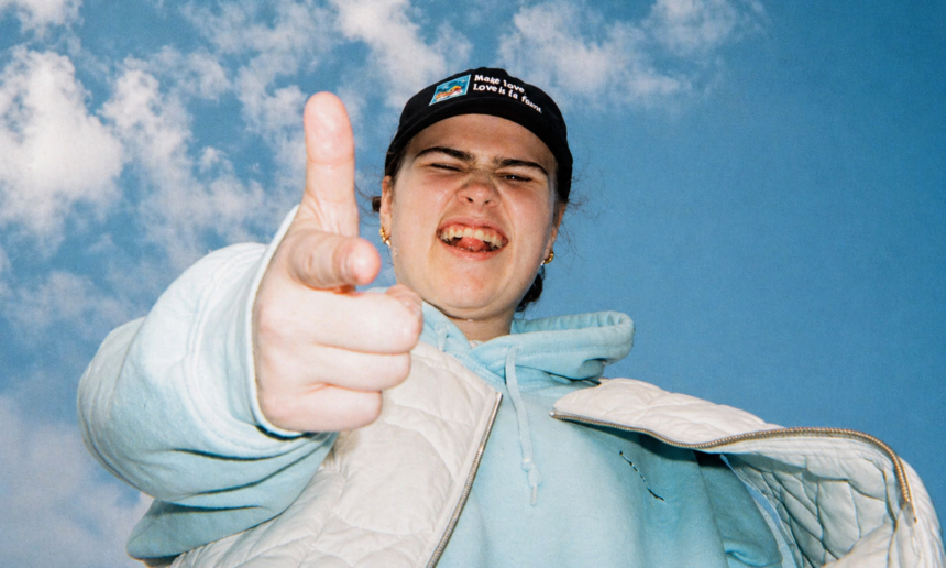 Venbee points playfully at the camera under a bright blue sky, wearing a black cap, light blue hoodie, and white jacket, capturing a candid, energetic moment.