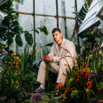 Wilkinson sits in a lush, surreal garden surrounded by reflective shards and vivid flowers, holding a red bloom, echoing the atmospheric intensity of “Infinity.”