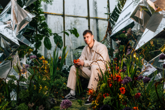 Wilkinson sits in a lush, surreal garden surrounded by reflective shards and vivid flowers, holding a red bloom, echoing the atmospheric intensity of “Infinity.”