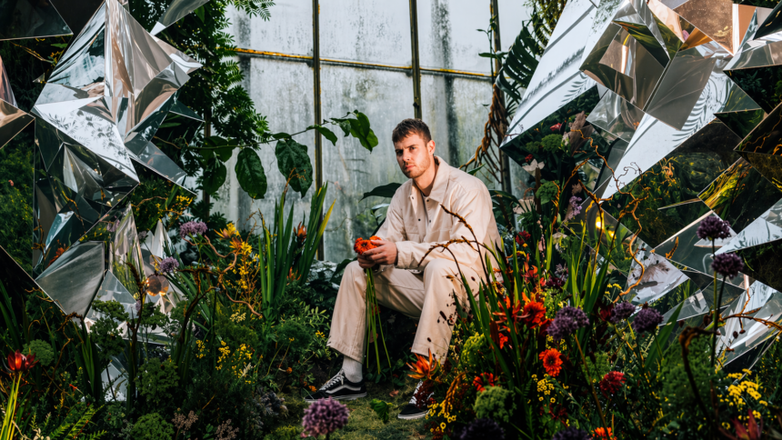 Wilkinson sits in a lush, surreal garden surrounded by reflective shards and vivid flowers, holding a red bloom, echoing the atmospheric intensity of “Infinity.”