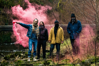 Wu Lyf band members wearing masks and standing in front of rocky terrain with pink smoke, promoting their 2026 UK and European tour
