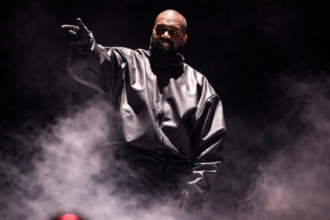 Ye performs on a darkened stage, wearing an oversized grey jacket and pointing toward the crowd, with stage smoke billowing around him.