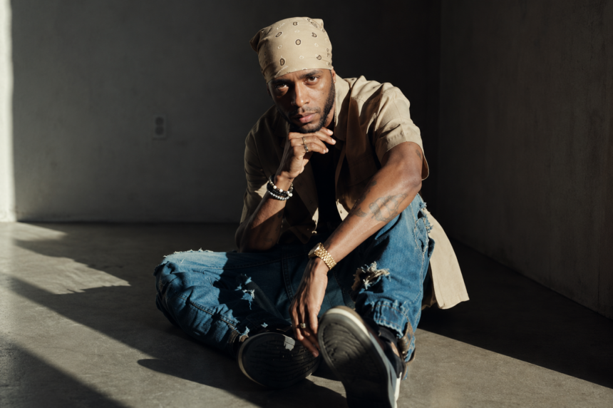 **Alt text:** 6LACK sits on the floor in a relaxed pose against a neutral studio backdrop, wearing a white bandana, beige shirt, and distressed jeans, in a press photo for his 2026 "10 Years of 6LACK" world tour.