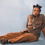 `Dua Saleh seated on the floor in a brown utility shirt and wide-leg trousers with a chain and padlock necklace, photographed against a soft blue studio backdrop.`
