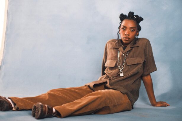 `Dua Saleh seated on the floor in a brown utility shirt and wide-leg trousers with a chain and padlock necklace, photographed against a soft blue studio backdrop.`
