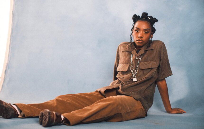 `Dua Saleh seated on the floor in a brown utility shirt and wide-leg trousers with a chain and padlock necklace, photographed against a soft blue studio backdrop.`