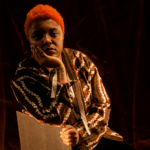 `Loraine James in a patterned shirt with vibrant orange hair, resting her chin on her hand in a moody, warmly lit portrait shoot.`