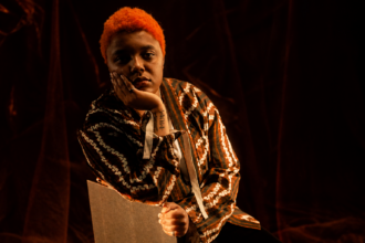 `Loraine James in a patterned shirt with vibrant orange hair, resting her chin on her hand in a moody, warmly lit portrait shoot.`