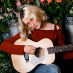 Maisie Peters playing acoustic guitar surrounded by flowers and flower buckets for her 2026 album 'Florescence' press shoot