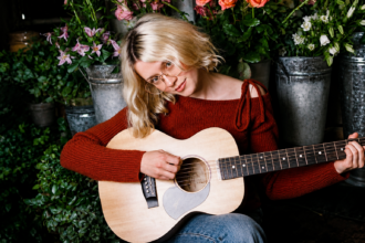 Maisie Peters playing acoustic guitar surrounded by flowers and flower buckets for her 2026 album 'Florescence' press shoot