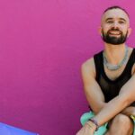 Olof Dreijer press photo against a pink wall, smiling in a black mesh top and layered jewelry