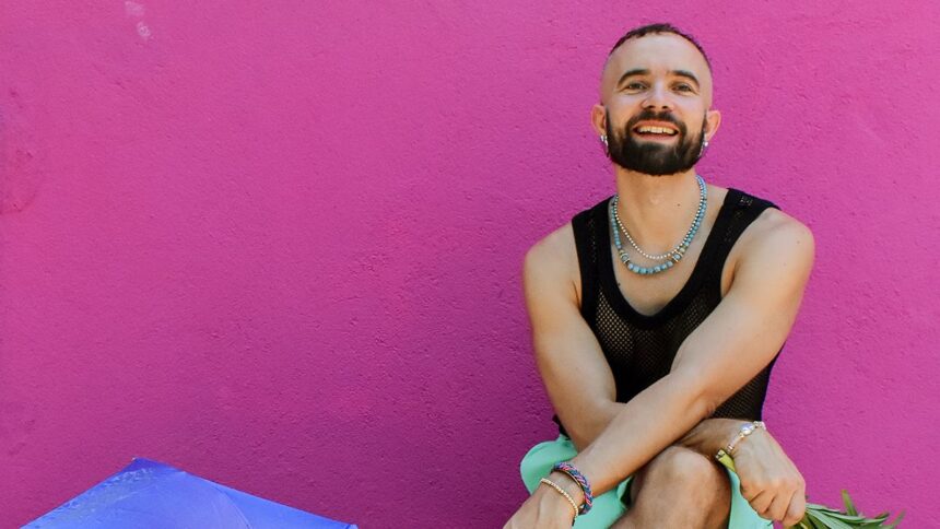 Olof Dreijer press photo against a pink wall, smiling in a black mesh top and layered jewelry