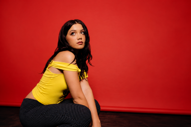 `Alessi Rose poses in a yellow off-shoulder top against a red backdrop in a press photo for her new single 'Skin.'`