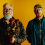 `Arab Strap's Aidan Moffat and Malcolm Middleton pose against a yellow backdrop, with Moffat holding a bunch of dried red roses, in a press photo for their 2026 album 'Half-Told Tales.'`