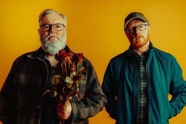 `Arab Strap's Aidan Moffat and Malcolm Middleton pose against a yellow backdrop, with Moffat holding a bunch of dried red roses, in a press photo for their 2026 album 'Half-Told Tales.'`