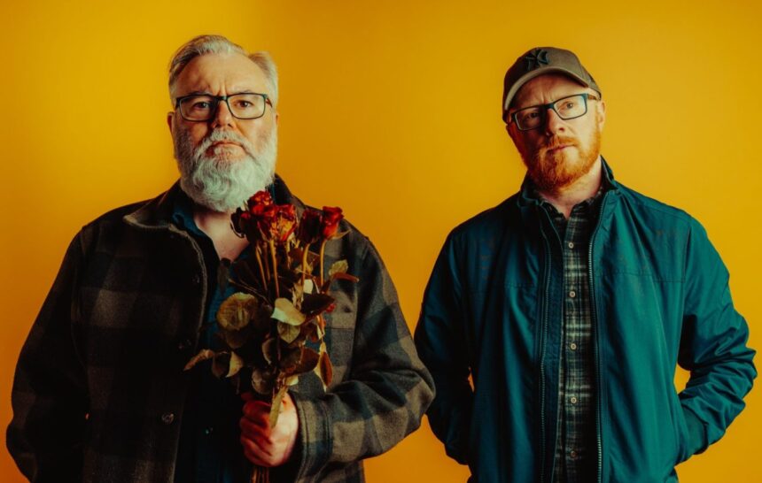`Arab Strap's Aidan Moffat and Malcolm Middleton pose against a yellow backdrop, with Moffat holding a bunch of dried red roses, in a press photo for their 2026 album 'Half-Told Tales.'`